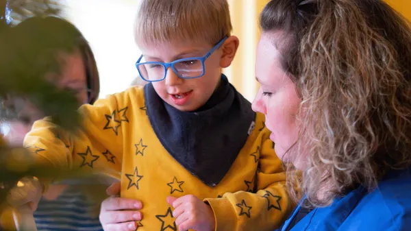 Ein Kind mit blauer Brille und einem gelben Pullover mit Sternenmuster lächelt, umgeben von zwei Erwachsenen, von denen einer ihm Halt gibt.
