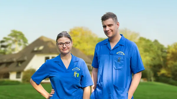 Zwei Personen in blauen Uniformen stehen draußen vor einem verschwommenen Haus und Bäumen im Hintergrund und lächeln in die Kamera.