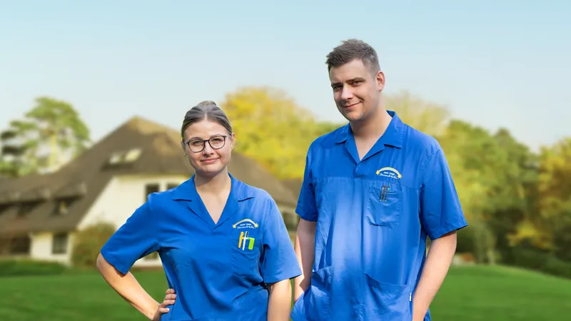 Zwei Personen in blauen Uniformen stehen draußen vor einem verschwommenen Haus und Bäumen im Hintergrund und lächeln in die Kamera.