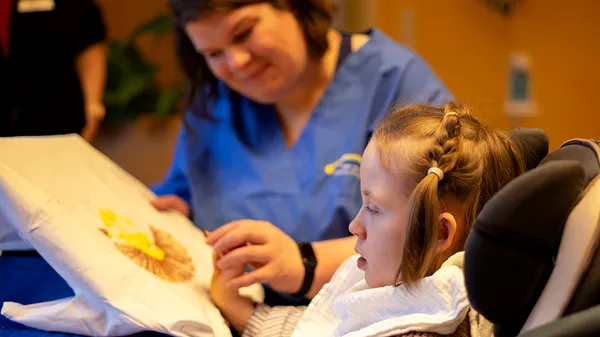 A young girl in a wheelchair paints with assistance from a woman in a blue uniform, focusing intently on the canvas.