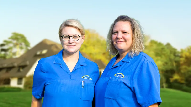 Zwei Frauen in blauen Uniformen stehen lächelnd im Freien vor einem verschwommenen Hintergrund aus Bäumen und einem Gebäude.