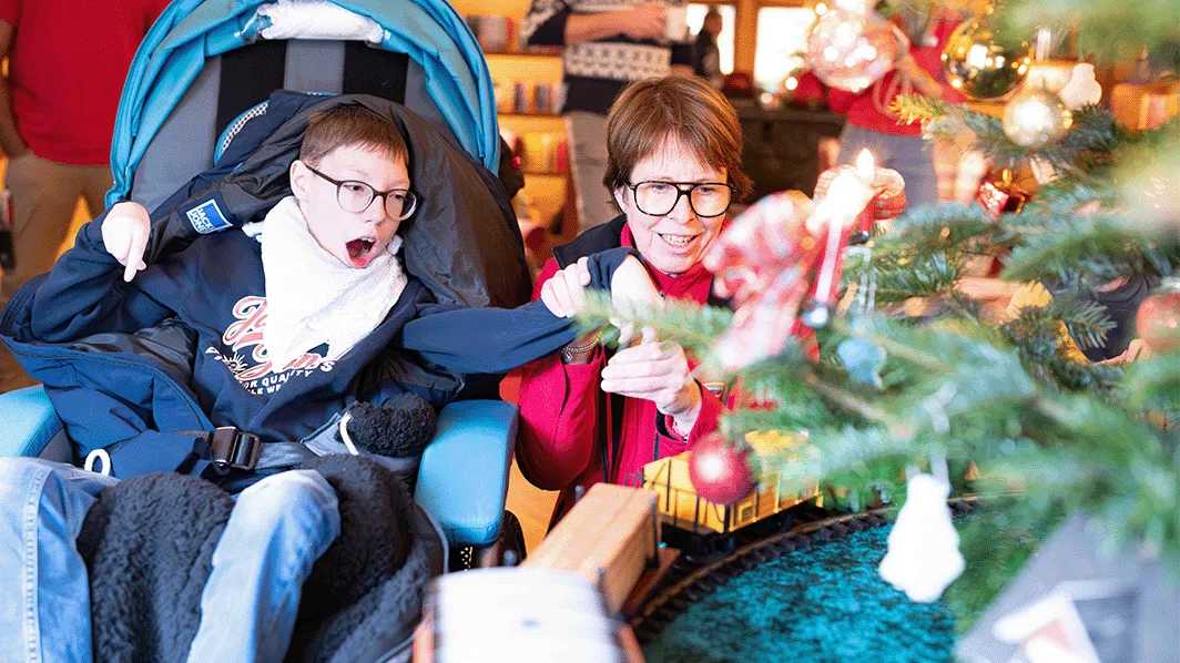 Eine Person im Rollstuhl und eine weitere Person beobachten voller Freude eine Modelleisenbahn, die um einen Weihnachtsbaum in einem warm beleuchteten Raum fährt.