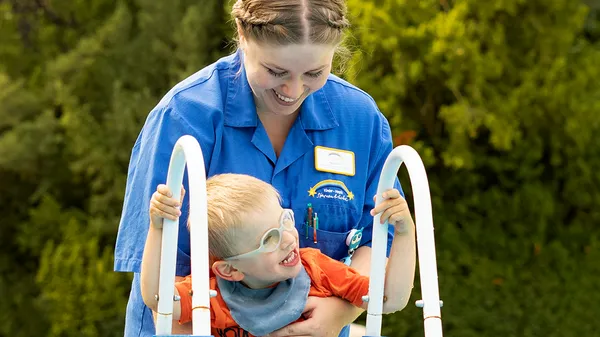 A woman in blue work clothes holds a little boy steady as he climbs a ladder. Both are smiling.