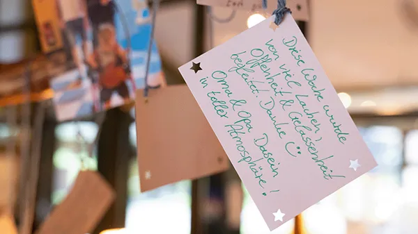 Eine handgeschriebene Notiz in deutscher Sprache hängt an einer Schnur, umgeben von verschwommenen Hintergrundelementen und sanfter Beleuchtung.