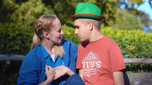 A woman in blue work clothes and a young teenager wearing a hat are sitting on a bench, holding hands.