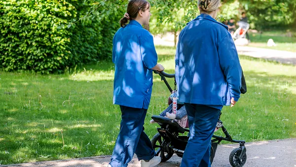 Zwei erwachsene Personen in blauer Berufskleidung schieben einen Kinderwagen durch einen Garten. Im Kinderwagen liegt ein Kind.