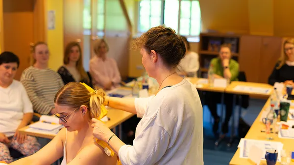 Several people are seated at tables in a seminar room. A woman is standing and placing a yellow towel around the neck of a seated woman.