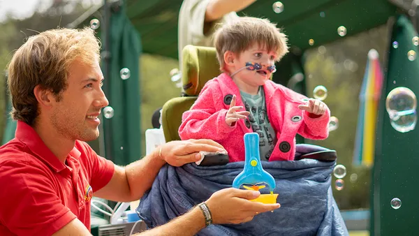 A young man in a red polo shirt and a small child in a wheelchair are blowing soap bubbles together. Several bubbles float in the air around them.