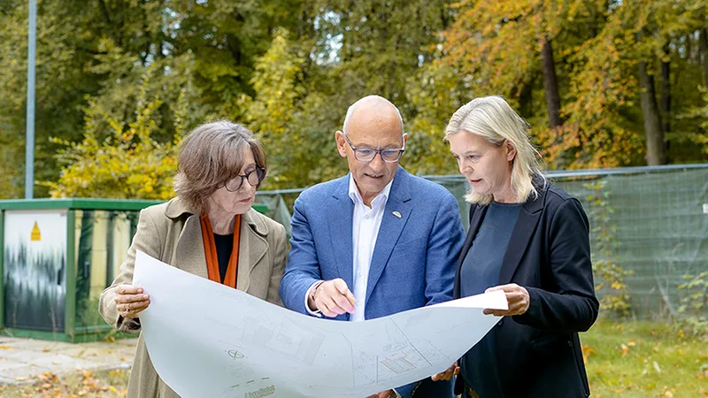 A man and two women are standing outside, discussing a large construction plan.