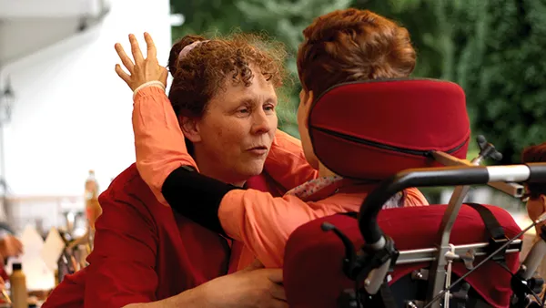 Eine Person in einem roten Hemd umarmt ein Kind im Rollstuhl, beide tragen orangefarbene Kleidung. Die Szene scheint im Freien zu spielen, mit viel Grün im Hintergrund.