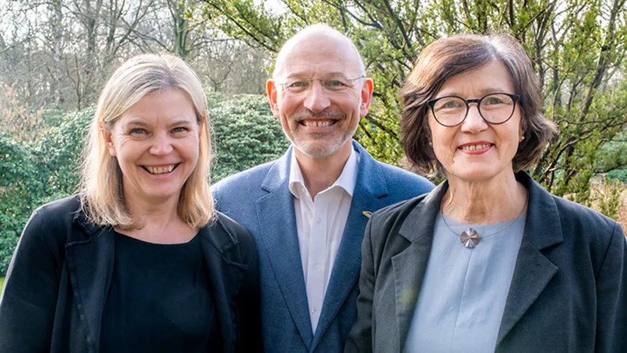 Three people smiling outdoors, with trees and shrubs in the background. The group includes two women and one man, all wearing business attire.