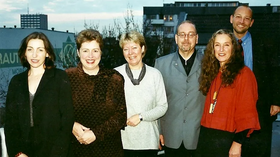Six people standing together outdoors, smiling, with buildings and a cloudy sky in the background.