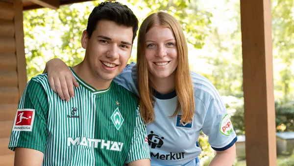 Ein junger Mann und eine junge Frau lächeln, tragen grüne und blaue Fußballtrikots und sitzen zusammen im Freien vor einem Hintergrund mit Bäumen.