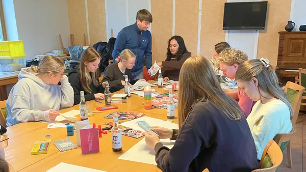 Eine Gruppe von Menschen sitzt um einen Tisch herum und beschäftigt sich mit Bastelarbeiten, wobei Kunstzubehör verstreut liegt und eine Person den anderen hilft.