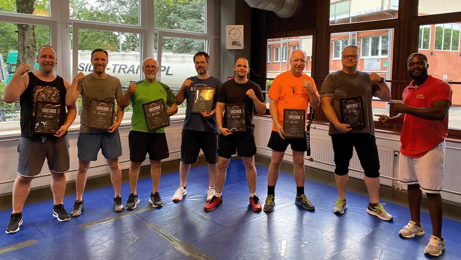 Acht Männer stehen in einem Fitnessstudio und halten Zertifikate, während sie selbstbewusst posieren. Sie wirken stolz und freuen sich.