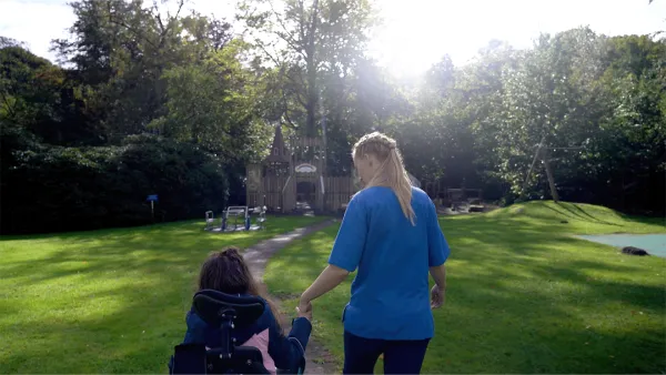 Eine Pflegekraft hält die Hand einer Person im Rollstuhl und spaziert mit ihr durch einen sonnendurchfluteten Park mit üppigem Grün und Bäumen.