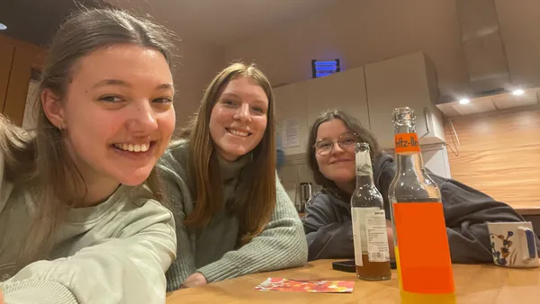 Drei Personen sitzen lächelnd an einem Tisch mit Getränken, darunter eine Flasche Orangensoda, in einem gemütlichen, warm beleuchteten Raum.