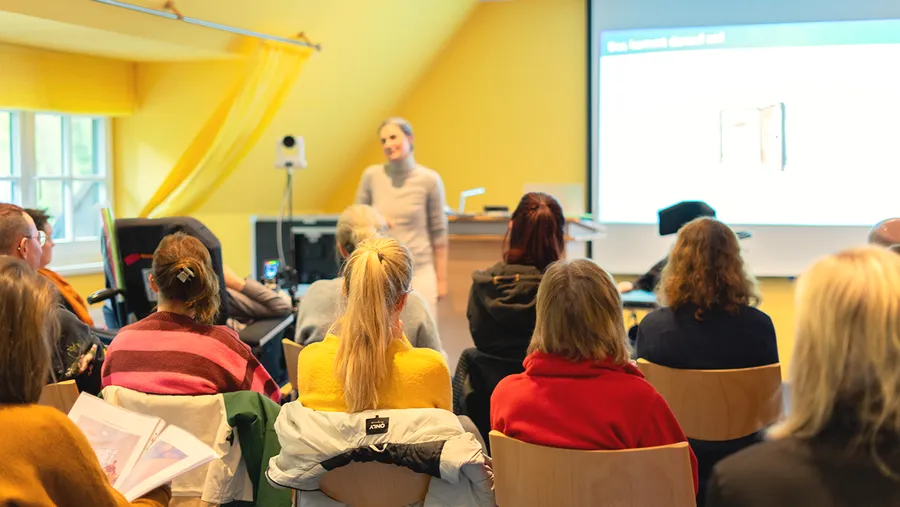 Eine Frau hält vor Publikum in einem hellen Raum mit gelben Wänden eine Präsentation. Im Hintergrund wird der Inhalt auf einer Projektionsleinwand angezeigt.
