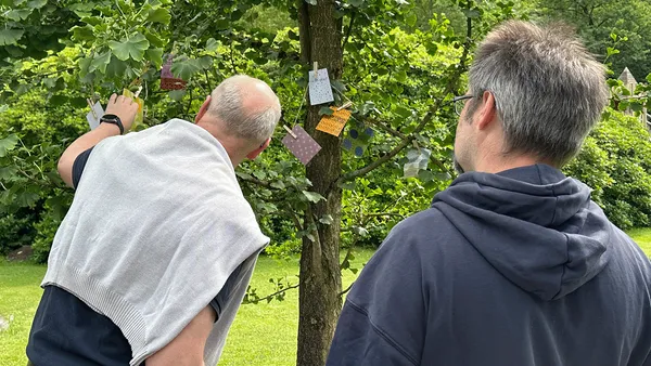 Zwei Männer betrachten einen Baum, an dem bunte Karten mit Wäscheklammern hängen, in einem grünen Garten.