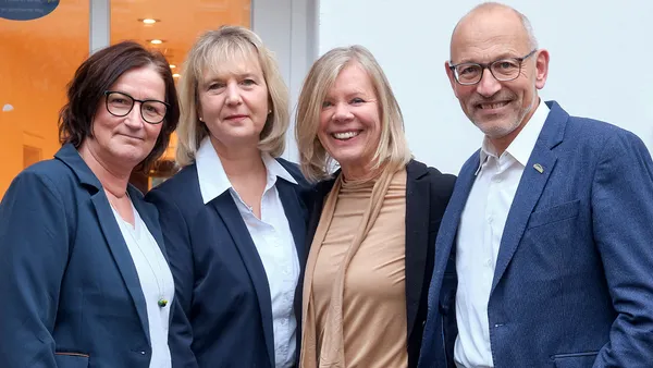 Vier Personen in Business-Kleidung stehen dicht beieinander und lächeln vor einer weißen Wand und einem beleuchteten Innenraum.