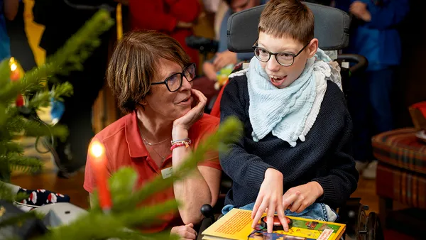 Ein fröhlich strahlender Junge im Rollstuhl und eine ihm zugewandte Frau im roten T-Shirt, sitzen neben einem Tannenbaum mit roten Kerzen.
