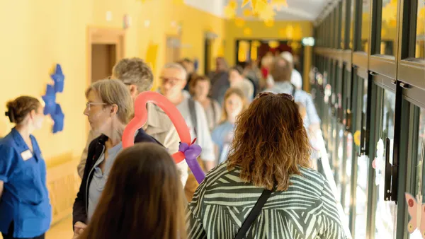 Menschen haben sich in einem hell erleuchteten Flur versammelt, der mit Papierfiguren geschmückt ist. Eine Person hält ein rosafarbenes Ballonherz.