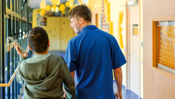 Eine Person in blauer Arbeitskleidung geht mit einer anderen Person, die ein Tablet in der Hand hält, durch einen hellen, mit gelben Sternen verzierten Flur.