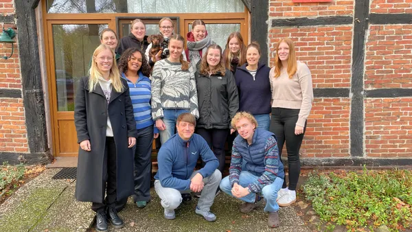 Eine Gruppe von Menschen, die draußen lächeln und auf einem Weg vor einem Backsteingebäude mit einer Holztür und Fenstern stehen. Freude & Vergnügen