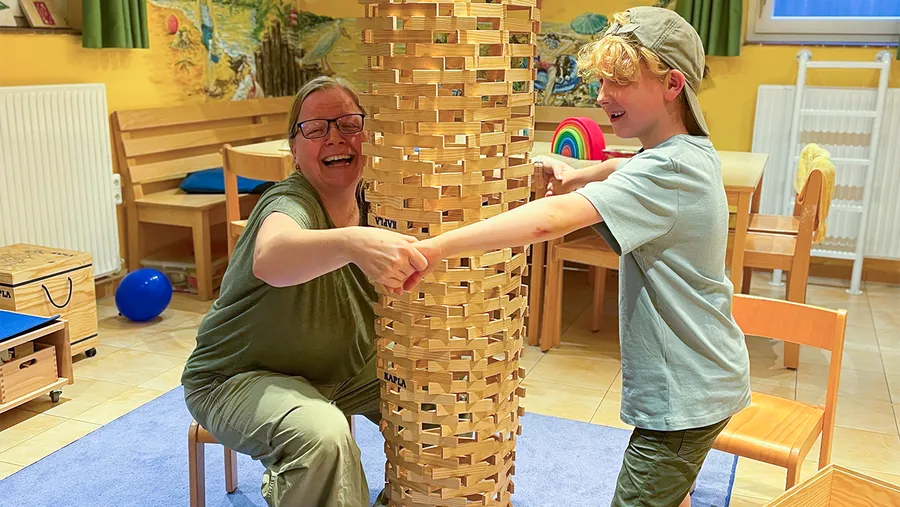 A smiling adult and child collaboratively build a tall tower from wooden blocks in a colorful room with chairs and a blue mat.