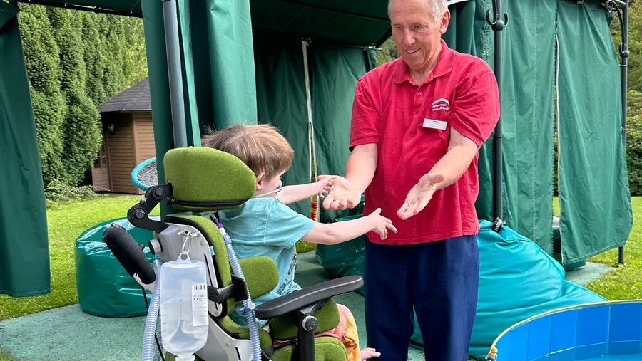 A man in a red shirt helps a child in a green wheelchair reach out, set outdoors with green structures and bean bags in the background.