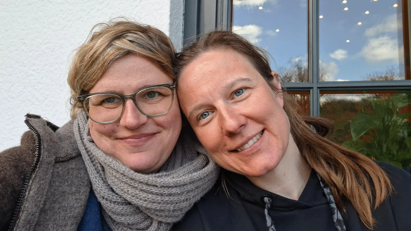 Zwei Frauen lächeln in die Kamera, eine trägt eine Brille und einen Schal, die andere hat einen Pferdeschwanz. Hintergrund: Fenster mit Bäumen.