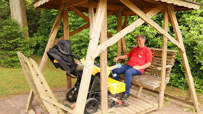 Man in red shirt sitting next to a pram in a wheelchair swing in a garden.