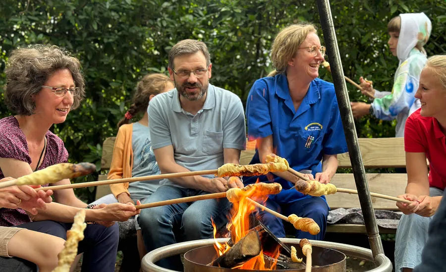 A group of people sitting around a campfire, smiling and roasting bread on sticks, with greenery in the background.