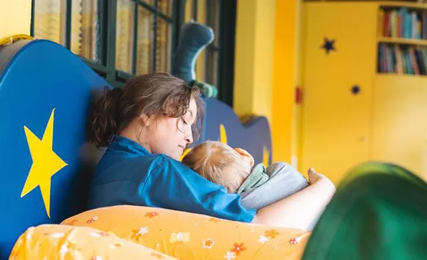 A woman gently cradles a sleeping child on a colorful, star-decorated bench in a cozy, yellow-walled room with bookshelves.