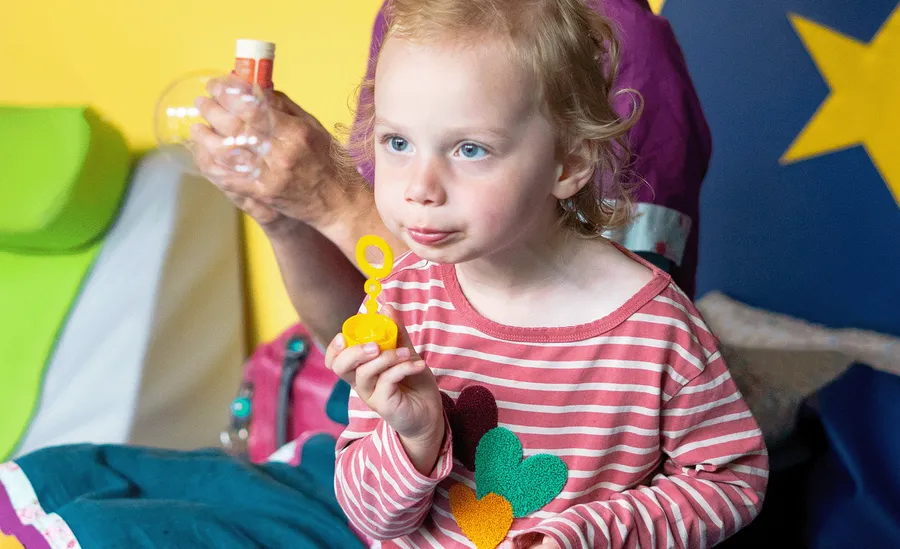 A young child with curly hair blows bubbles, wearing a pink striped shirt with colorful heart shapes. A person in the background holds a bubble bottle.