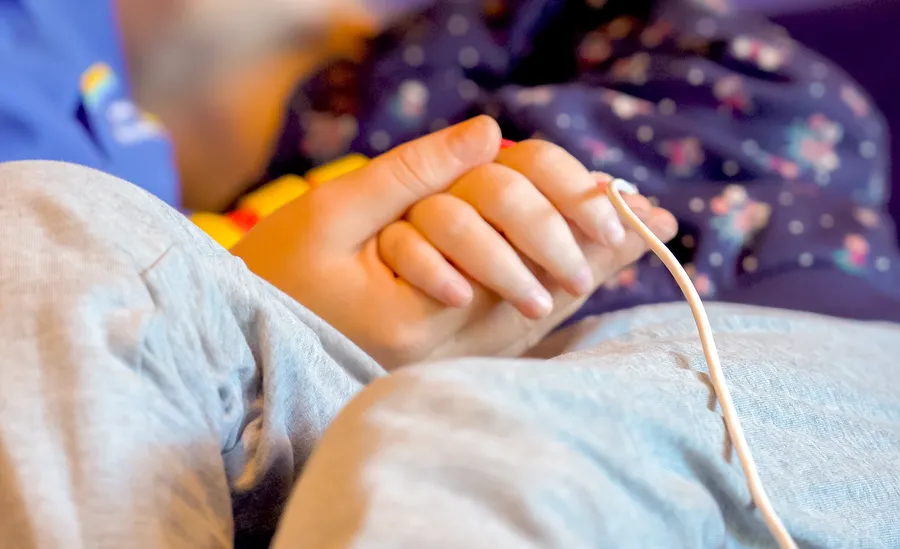 Die Hand einer erwachsenen Person hält die Hand eines Kindes. Ein medizinischer Schlauch liegt daneben und führt zum Kind.