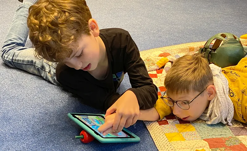 Two boys lying on a quilted blanket, focused on a tablet. One points at the screen, both engaged in the activity.