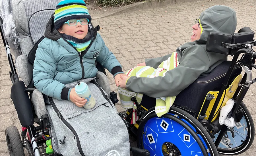 Zwei Kinder im Rollstuhl halten sich an den Händen. Das eine trägt eine Brille und einen gestreiften Hut und hält eine Flasche in der Hand. Das andere ist in eine bunte Decke gehüllt.