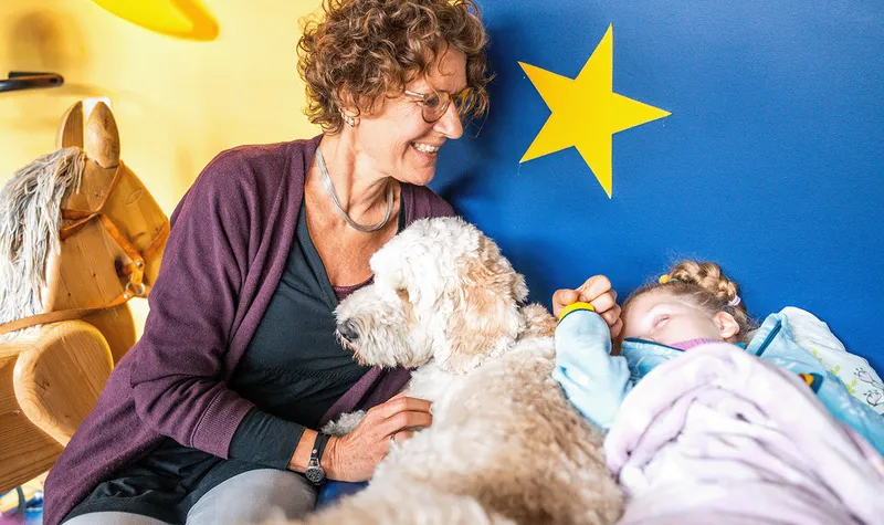 A woman with curly hair gently holds the hand of a sleeping girl. A dog lies between them.