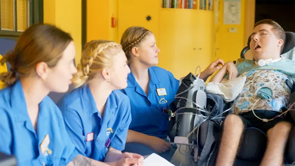Drei Pflegekräfte in blauen Uniformen sitzen da und beobachten aufmerksam einen jungen Menschen mit einem medizinischen Gerät in einem Rollstuhl in einem hellen Raum.