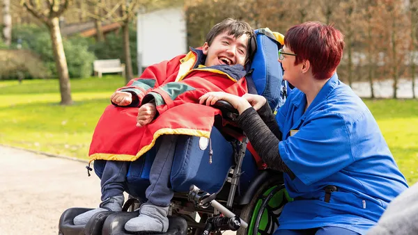 A woman in blue work clothing is kneeling next to a young man in a wheelchair. The young man is wearing a red blanket and laughing while the woman smiles at him.