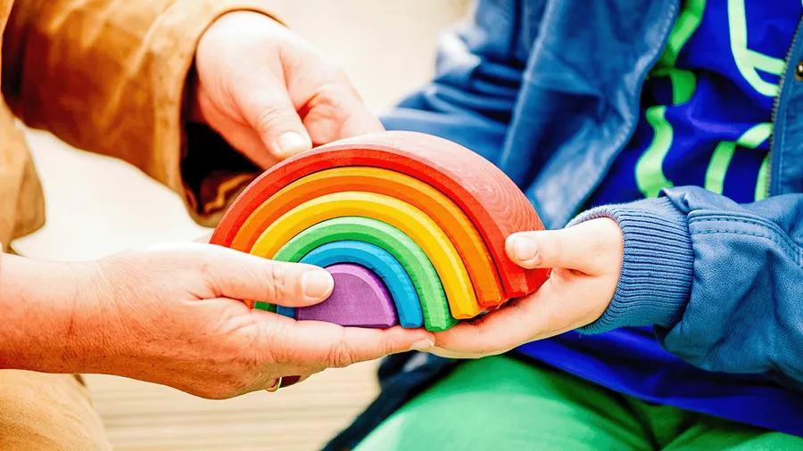 Die Hände einer erwachsenen Person und die Hände eines Kindes halten zusammen einen bunten, hölzernen Regenbogen, bestehend aus mehreren Teilen.