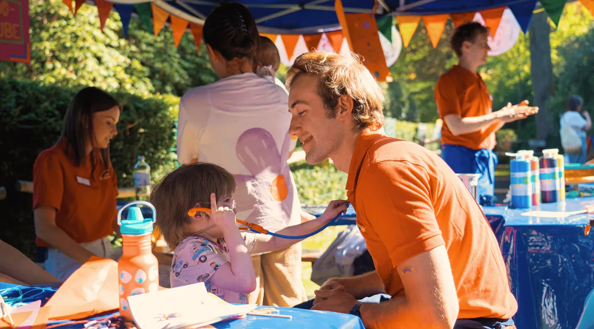 Ein Kind untersucht spielerisch das Gesicht eines lächelnden Mannes mit einem Stethoskop an einem Marktstand im Freien. Bunte Dekorationen und Verkaufsartikel umgeben sie.