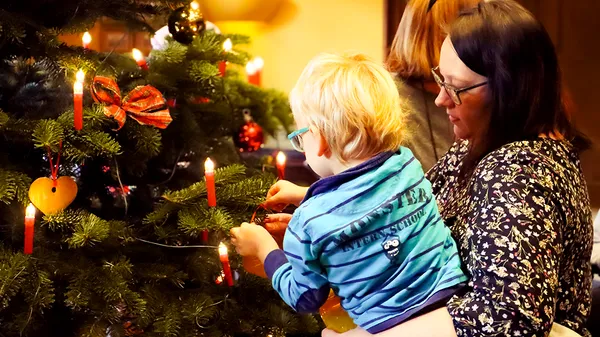 Eine Frau hält ein Kind im Arm, während sie einen Weihnachtsbaum mit roten Kerzen und Christbaumschmuck schmücken. Ein weiteres Kind sitzt in der Nähe und trägt einen festlichen Pullover.