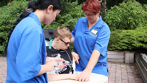 Two caregivers in blue uniforms assist a young person in sunglasses, seated outdoors, with a medical procedure.