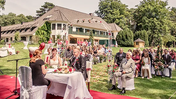 Outdoor wedding ceremony with a couple seated at a table, guests watching, and a large house in the background surrounded by trees.