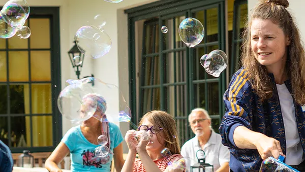 A woman and a child joyfully play with bubbles outdoors, surrounded by a few seated adults.