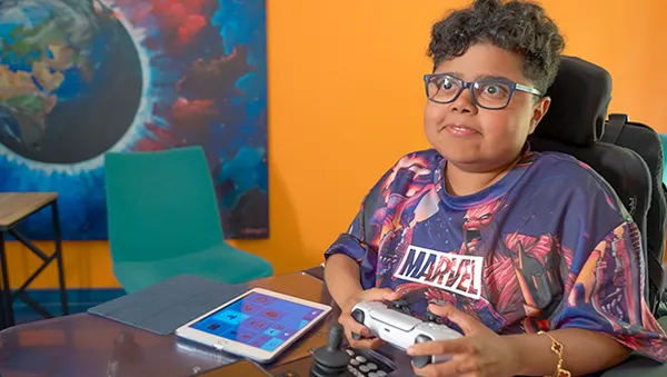 A person in a Marvel shirt plays with a game controller, sitting in front of a tablet. A colorful planet painting is in the background.