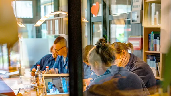 People in a meeting room, some looking at a laptop screen. The room has bookshelves, and a heart-shaped decoration is visible on the window.