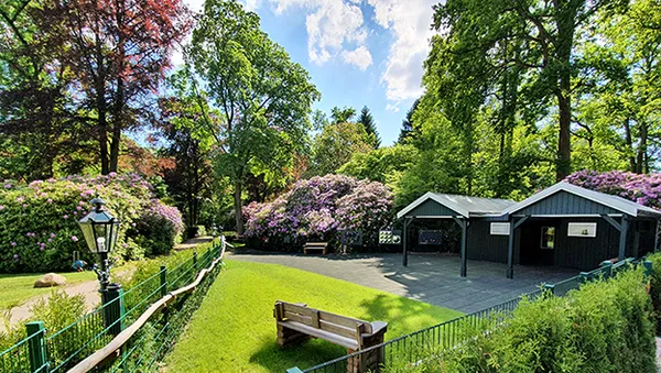 A lush garden scene with vibrant flowers, a wooden bench, a lamp post, and two small black buildings under a bright blue sky.